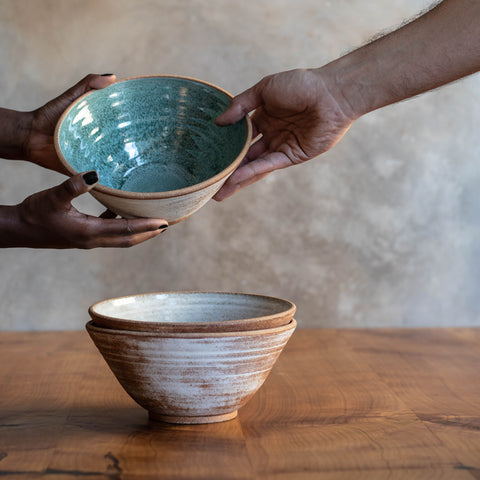 A stack of two Ramen Bowls on a wooden table. A hand passing a Ramen Bowl with Thai Blue glaze to a another's hands.