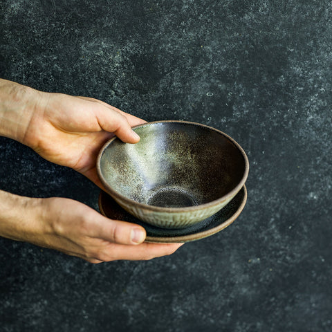 Hand holding a Progress Cereal Bowl glazed in Eelskin, displayed with a Cafe Plate