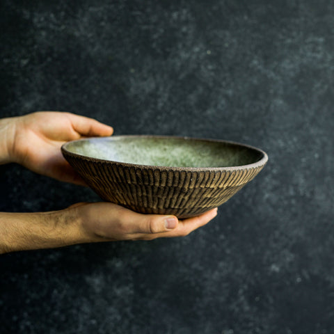 Hand presenting a Progress Serving Bowl, glazed in Moss Green. The dramatic lighting highlights the hand etched texture, glazed in Brown Leather Matte