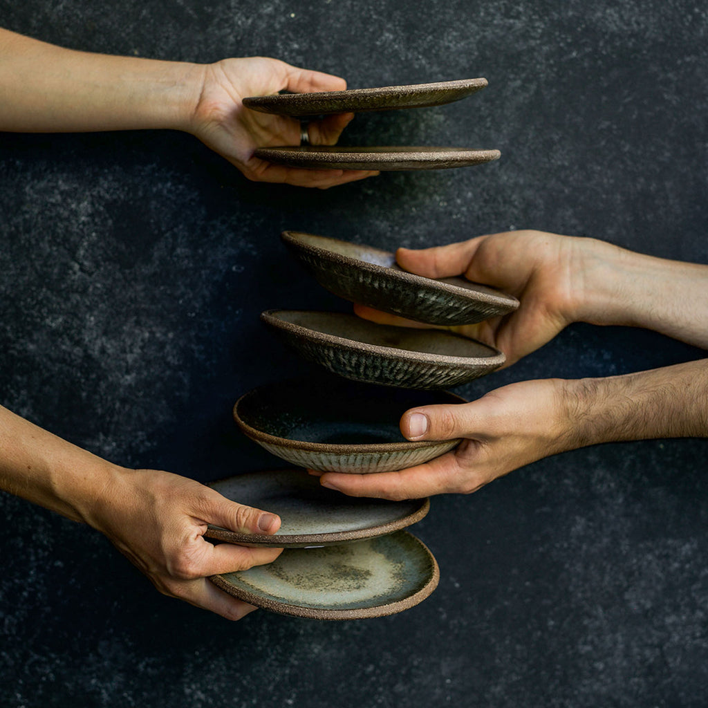 Hands holding stacks of Progress Cafe Plates and Dessert Bowls, in front of a dark gray backdrop