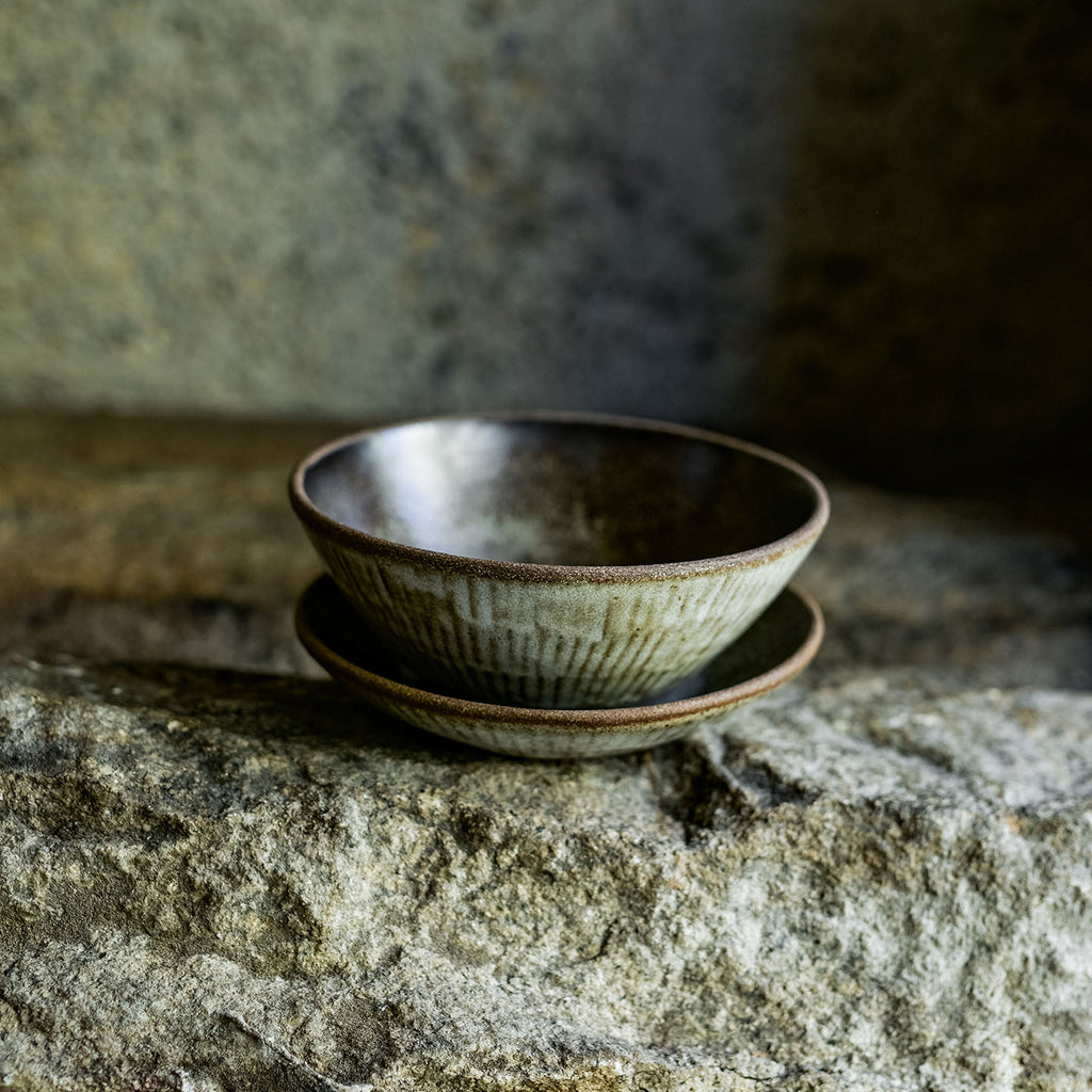 Progress Cereal Bowl glazed in Eelskin, displayed with a Cafe Plate, in dramatic lighting on a rocky textured surface