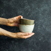 A hand holding two Progress Rice Bowls, with exterior glazes of Moonshadow and White Chamois