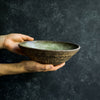 Hand presenting a Progress Serving Bowl, glazed in Moss Green. The dramatic lighting highlights the hand etched texture, glazed in Brown Leather Matte