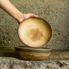 Hand holding a Brown Leather Matte Progress Stu Bowl, exposing its interior, above a rocky earthy surface